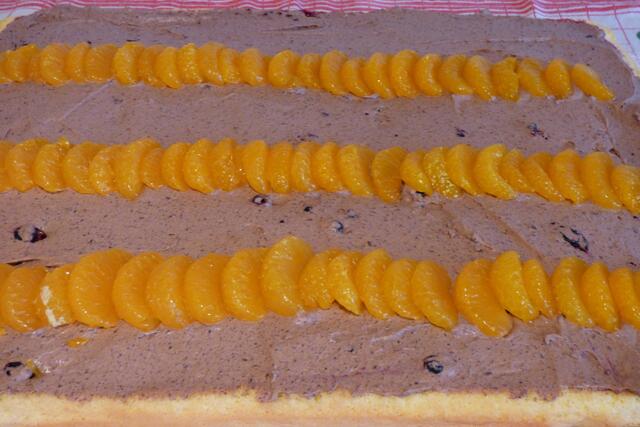 Die aufgeschlagene Schokosahne gleichmäßig aufstreichen. Auch das geht wieder am besten mit der Kuchenpalette. Dann bspw. Mandarinenschnitze oder anderes Obst (Himbeeren, Erdbeeren, Blaubeeren, Bananen, Aprikosenstücken etc.) darauf verteilen.