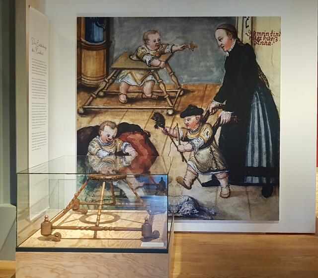 Sehr schön ist hier die Präsentation eines historischen Geh- / Gängelwagens (im Glaskasten) vor einer historischen Illustration, links im Bild der Überblickstext (den ich leider nicht gesondert fotografiert habe).

Der Bewegungsradius von Kleinkindern war in früheren Zeiten im Vergleich zu heute erheblich eingeschränkt. Allerdings kann ich mich noch an Laufgurte und Gehwagen in den 1950er Jahren erinnern. | Foto: eigene Aufnahme 