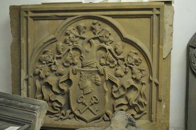 Wappenstein Firma Zwirnerei Ackermann Sontheim. ursprünglich befand sich der Stein am Pförtnerhaus, das an die Kolpingstraße 4 (ehem. Sommervilla des Deutschordenskomturs) angebaut war. Das Pförtnerhaus wurde 1960 durch einen großen Neubau abgelöst. 
Der Stein zeigt als Wappen einen Frauenkopf und gekreuzte Schlüssel (dieses Markenzeichen ist ab 1895 nachweisbar). Der Wappenschild ist mit einem Band an eine Eiche mit Wurzeln und Astwerk gebunden.
Quelle: Stadtarchiv
 | Foto: Daniela Somers