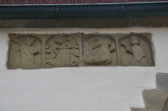 Ursprünglich war diese Steintafel nicht an der Außenwand der Nordseite. Von links nach rechts zeigen die Figuren: Einen stehenden Bischof mit Stab, den Erzengel Michael mit Drache, einen knienden Mönch mit Kapuze der etwas entgegen nimmt die ihm die Figur ganz rechts reicht. | Foto: Daniela Somers