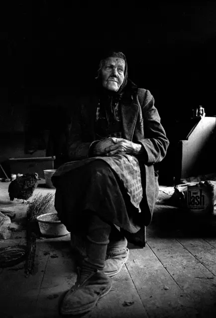 Pauline Schaffitzel, 
Einsiedlerin in der Mittelmühle

Roland Bauer erzählt:

Als ich im Dorf Ettenhausen nach der Mittelmühle fragte, hieß es nur: „Meinen Sie die Einsiedlerin?"- Pauline Schaffitzel sei früher das hübscheste Mädchen im Ort gewesen. Doch keiner war ihr recht, und so blieb sie ledig.

Der Weg führte mich zu einem verfallenen Gebäude am Ortsrand. Ich klopfte, rief - nichts rührte sich. Schon im Begriff zu gehen, öffnete sich plötzlich die Tür. Eine gebeugte alte Frau trat hervor, skeptisch, aber nicht abweisend. Nach kurzem Gespräch bat sie mich in ihr Haus.

Im Zentrum der dunklen Stube stand ein großer guss-eiserner Ofen. Alles war verrußt, der Raum von einem grauen Schleier überzogen. Pauline war fast blind. Sie lebte allein, mit ein paar Hühnern und kranken Katzen. An diesem Tag fotografierte ich nicht, ich hörte einfach zu.

Sie war das jüngste von drei Geschwistern. Der ältere Bruder fiel im Krieg, der jüngere hielt die Mühle noch einige Jahre am Laufen. Damals mahlte das Wasserrad fast rund um die Uhr. Später verdrängten industrielle Mühlen den kleinen Betrieb. Nach dem Tod des Bruders blieb Pauline allein zurück. Die Mühle verfiel, das Dorf wandte sich ab.

Mit über 80 Jahren hatte sie dann nur noch eine Kuh, kaum Milch, keine Krankenversicherung. Nach einem Oberschenkelbruch konnte sie sich nur noch mit einem Holzschemel fortbewegen.

An Weihnachten saß sie, in Decken gehüllt, auf dem Sofa. Es war kalt, kein Licht, kaum Wärme. Wir waren die Einzigen, die sie besuchten. Zum Abschied schenkte sie uns eine vergilbte Packung Kekse - es waren die, die wir ihr einst mitgebracht hatten. | Foto: Foto und Text: Roland Bauer