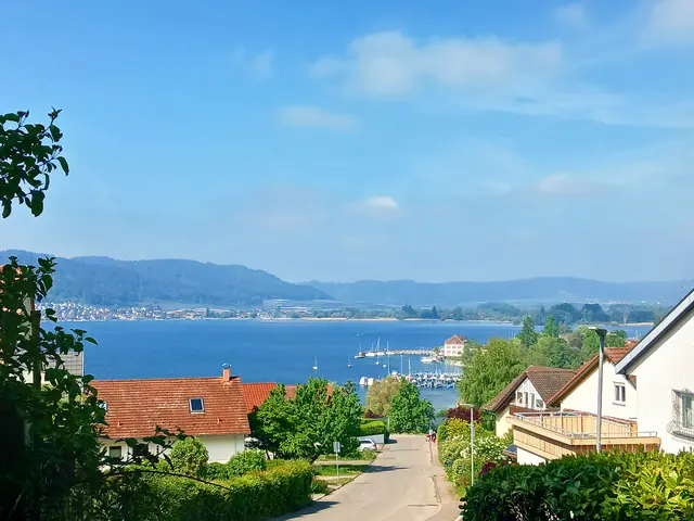 Man schaut hinunter auf den blauen Bodensee, rechts und links stehen Häuser,  in der Mitte verläuft die Straße zum See.  Auf der anderen,  weit entfernten Uferseite sieht man eine grüne Hügelkette.  