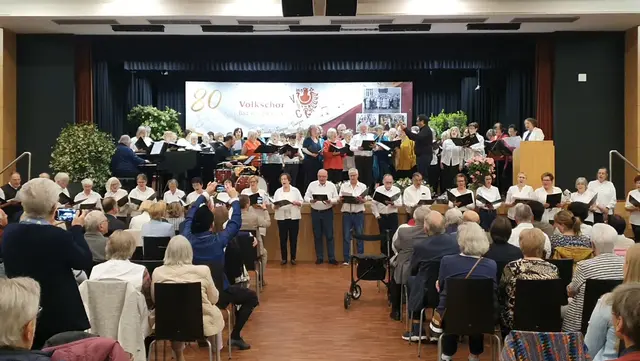 Vioele Menschen vor und auf der Bühne mit Noten in der Hand, die gemeinsam singen im Vordergrund viele Zuschauer