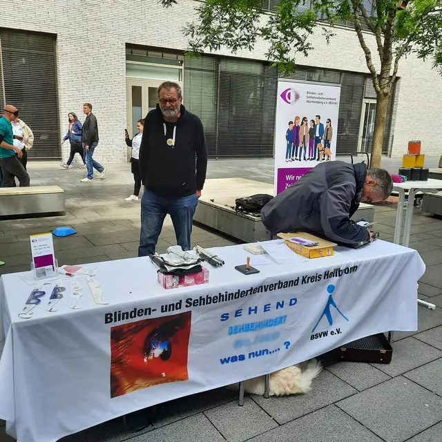 Infostand mit zwei Männern hinter einem Tisch mit Banner. Der Tisch steht im Freien auf einem gepflasterten Platz, im Hintergrund sind ein modernes Gebäude mit großen Fenstern, einige Passanten und ein Baum zu erkennen. Hinter dem Tisch steht links ein Mann mittleren Alters mit grauen Haaren und Bart, hellem Hautton, schwarzem Kapuzenpullover und Jeans, die Hände in den Hosentaschen. Rechts beugt sich ein Mann mit grauen Haaren und Brille über den Tisch; er trägt eine dunkle Jacke. Auf dem Tisch liegen verschiedene Gegenstände, zum Beispiel Broschüren, ein paar kleine Hilfsmittel, eine Schachtel und weitere Kleinteile. Vor dem Tisch hängt ein weißes Banner mit mehreren Logos und Text. Darauf steht unter anderem gut lesbar: “Blinden- und Sehbehindertenverband Kreis Heilbronn” Darunter sind Logos und Texte wie “SEHEND” und “BSVH” sowie ein roter Bereich mit einer stilisierten Figur und dem Text “was nun…?” zu erkennen. Rechts hinter dem Tisch steht ein Roll-up-Banner mit lila Überschrift und einer Gruppe gezeichneter Personen; der Text ist teilweise zu erkennen, oben steht: “Blinden- und Sehbehindertenverband Württemberg e.V.” und darunter “Wir machen …” – der restliche Text ist unscharf.
