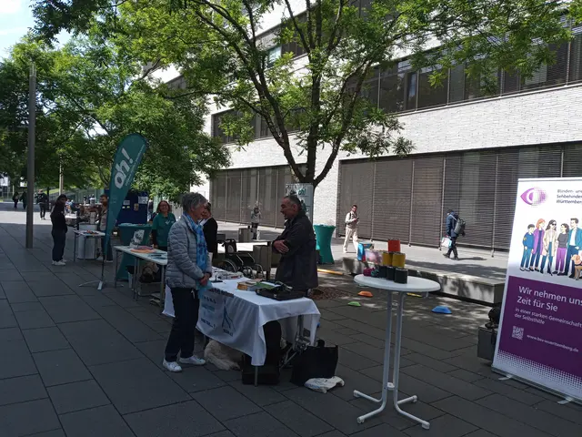 Straßenszene mit mehreren Stehtischen und Infoständen, offenbar bei einem Informations- oder Aktionstag im Freien. Menschen stehen an den Tischen, teils mit Jacken, und unterhalten sich oder schauen auf die ausgelegten Materialien. Links im Bild stehen mehrere Tische hintereinander auf einem gepflasterten Gehweg. Weiter hinten ragt eine grüne, hochstehende Beachflag mit der weißen Aufschrift „oh!“ empor. Rechts im Bild stehen Bäume mit grünen Blättern, dahinter ein modernes Gebäude mit hellen Fassaden und vielen Fenstern. Im unteren Bereich ist ein aufgestelltes, lila Werbeschild mit einer Gruppe gezeichneter Menschen und etwas Text zu sehen; dessen Text jedoch nicht klar lesbar. 