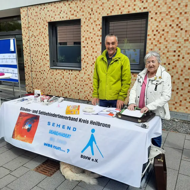Zu sehen ist ein Informationsstand im Freien. Er steht vor einer hell gemusterten Ziegelfassade und ist mit einem weißen Tuch bedeckt ist. Hinter dem Tisch stehen zwei Personen, eine Person links trägt eine leuchtend grüne Jacke und hat kurze graue Haare, die Person rechts hat graue, leicht lockige Haare und trägt eine helle Jacke über einem rosafarbenen Oberteil. Beide lächeln. Auf dem Tisch liegen verschiedene Gegenstände: mehrere Blätter Papier, ein paar Broschüren, ein orangefarbenes Päckchen, ein paar kleine Hilfsmittel sowie ein schwarzer Koffer. Vor dem Tisch hängt ein großes Banner mit Aufdruck. Der gut lesbare Text auf dem Banner: „Blinden- und Sehbehindertenverband Kreis Heilbronn“ Darunter befindet sich links ein Bild und Text, der verschwommen ist, aber teilweise lesbar: „SEHEND sehend werden was nun…?“ Rechts unten steht: „BSVW e.V.“ Unter dem Tisch schaut unten mittig das hellbeige Fell eines liegenden Hundes hervor, der sonst nicht zu erkennen ist. 