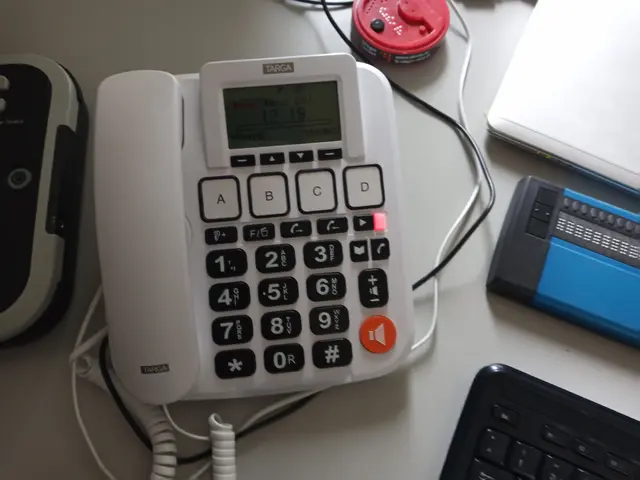 Gezeigt wird ein weißes Festnetztelefon mit sehr großen schwarzen Tasten auf einem Tisch. Oben am Telefon ist ein kleines Display; die Anzeige ist nicht lesbar. Direkt unter dem Display befinden sich vier rechteckige Kurzwahltasten mit der Beschriftung „A“, „B“, „C“ und „D“. Darunter ist ein Block mit großen, kontrastreichen Tasten: links von oben nach unten „1“, „4“, „7“, „*“, rechts daneben „2“, „5“, „8“, „0“, dann „3“, „6“, „9“ und eine Taste mit einem Pfeilsymbol. Rechts außen gibt es eine senkrechte Reihe mit Sondertasten, unter anderem eine mit einem Hörer‑Symbol und eine rote Taste mit einem Lautsprecher‑Symbol. Unten am Telefon gehen zwei Spiralkabel weg.