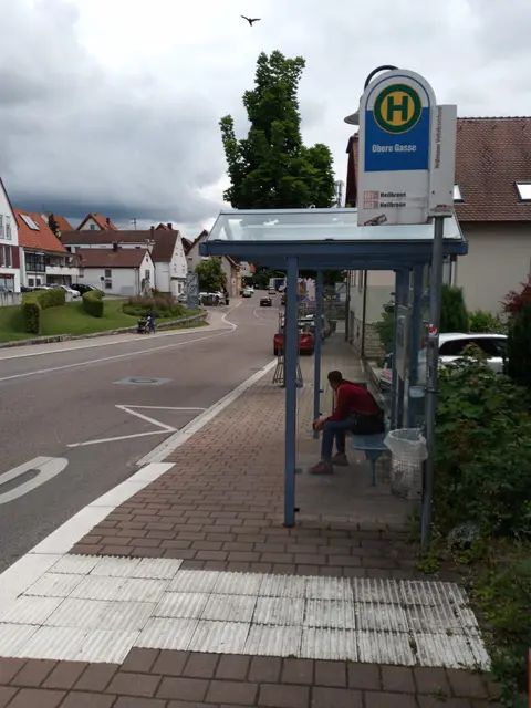 Bushaltestelle mit Wartehäuschen. Das Bild zeigt eine Straße mit Gehweg, Bushaltestelle und Häusern mit roten Dächern. Am rechten Bildrand steht das überdachte Wartehäuschen; darunter sitzt eine Person im roten Oberteil und dunkler Hose auf einer Bank. Daneben hängt ein weißer Mülleimer. Oben an einem Pfosten ist das grün-gelbe Haltestellenschild mit großem H zu sehen, darunter ein blauer Bereich mit der Aufschrift „Obere Gasse“. Die Straße verläuft leicht geschwungen nach links, rechts davon der gepflasterte Gehweg mit hellen, taktilen Bodenplatten im Bereich der Haltestelle. Im Hintergrund stehen weitere Wohnhäuser mit Gärten und ein großer Baum mit dichter Krone. Über allem ein bedeckter Himmel mit dunkleren Wolken. 